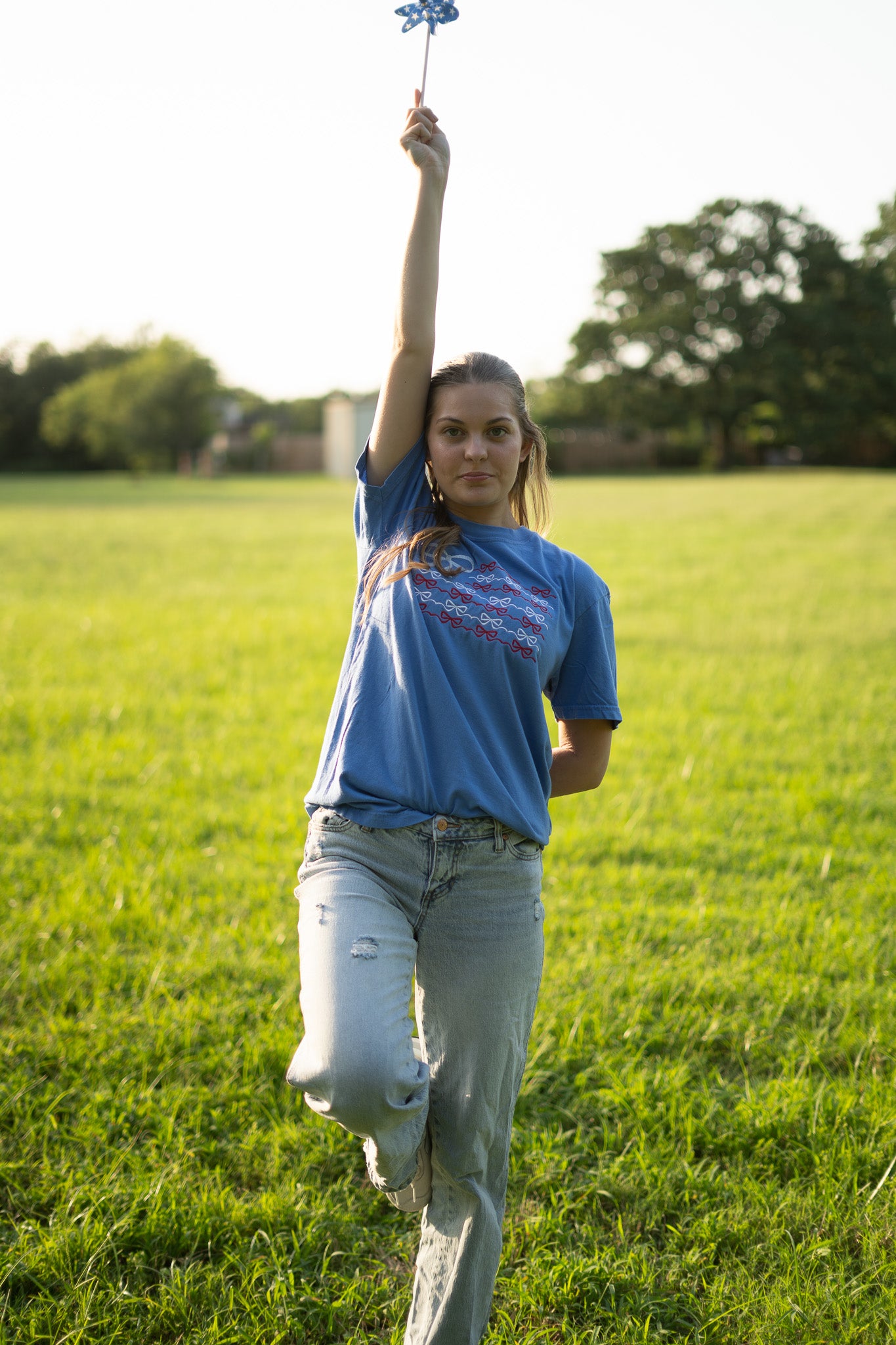 A young woman stands in a grassy field, holding a small blue pinwheel in her hand.