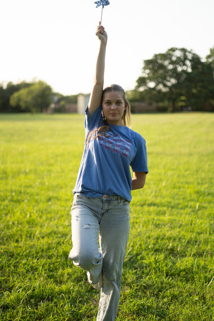 A young woman stands in a grassy field, holding a small blue pinwheel in her hand.